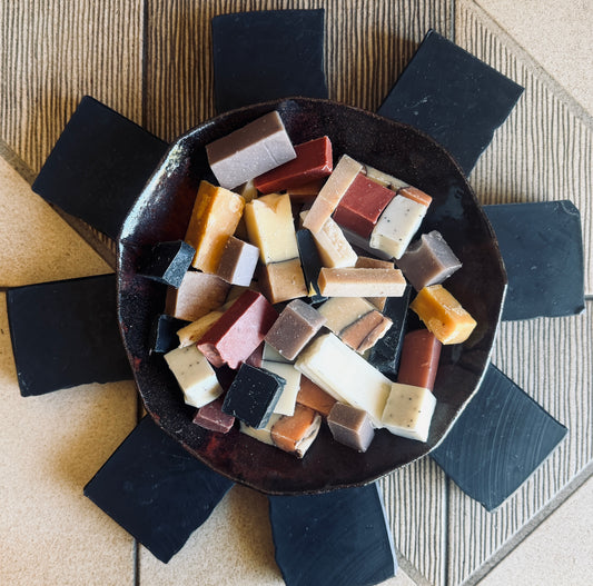 An overhead view of assorted rectangular bars of soap in various colors, including cream, orange, red, brown, and black, arranged in a dark ceramic bowl. The bowl is placed on a textured beige mat with several black bars of soap arranged around it.