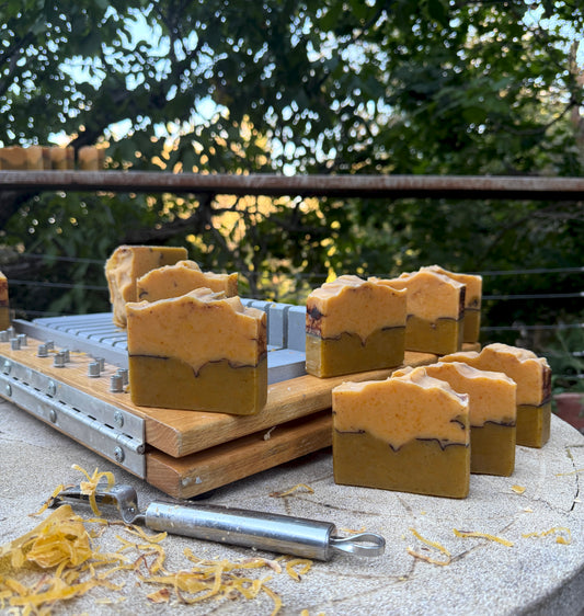 Several rectangular bars of handmade soap in warm tan and orange-brown layers are arranged on a wooden soap slicer outdoors on a stone table, with a metal tool and shredded yellow soap in the foreground and blurred greenery behind.