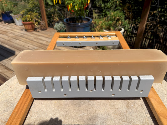 A tan rectangular bar of soap resting on a  soap slicer. Deck and potted plants are blurry in the background