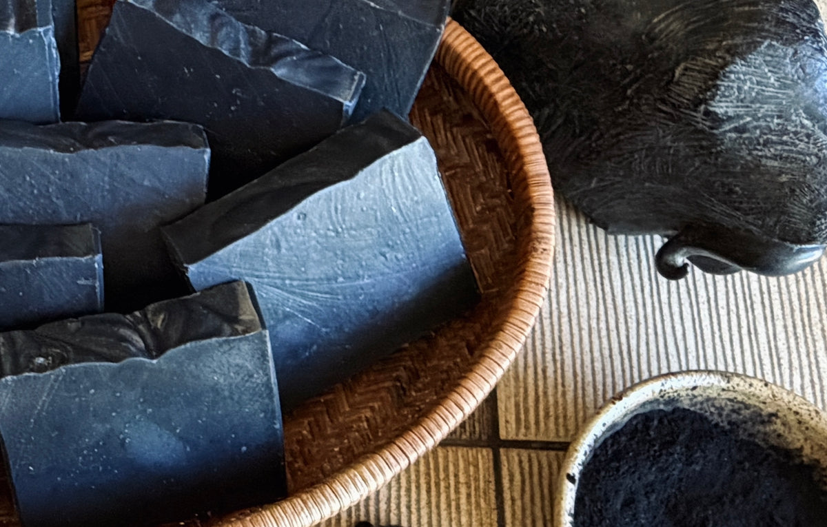 Dark charcoal-colored soap bars arranged in a woven basket, with a matching dark bowl and a textured black object on a striped mat background.