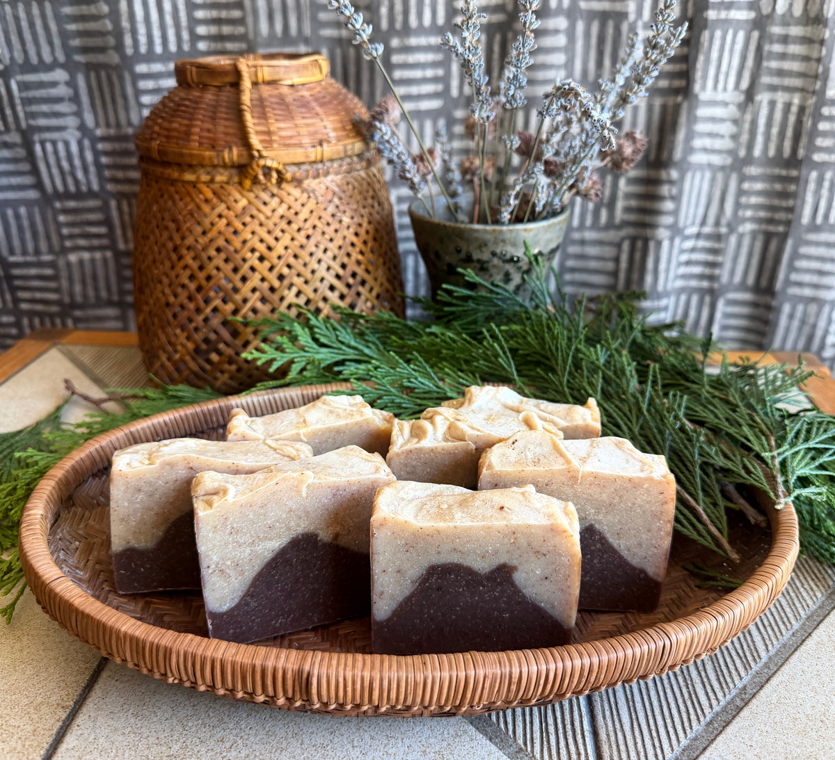 Several rectangular bars of layered soap in beige and dark brown sit on a round woven tray, surrounded by greenery, with a woven basket and patterned backdrop in the background.