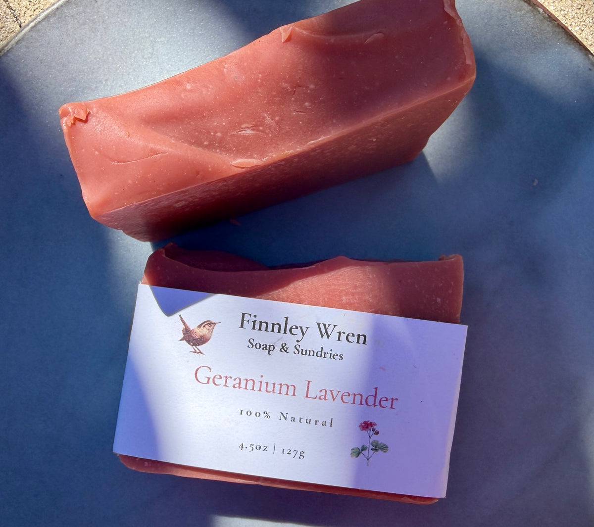 Two bars of Finney Wren Geranium Lavender soap on a slate blue background, with one bar featuring a white paper label.