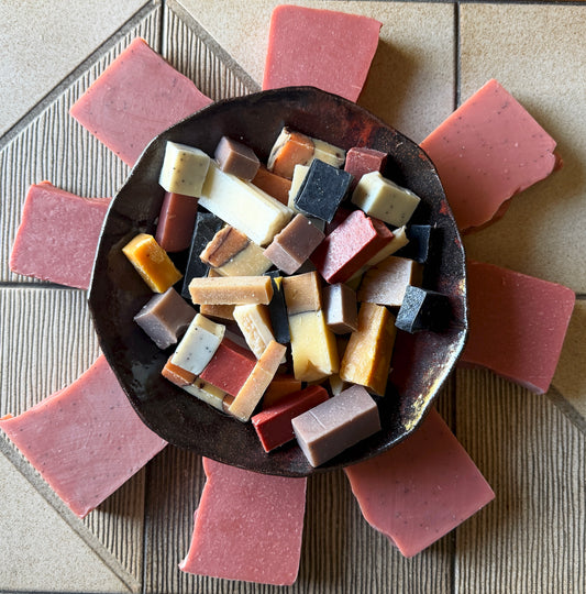 A top-down view of a rustic dark metal bowl filled with assorted rectangular bars of handmade soap, surrounded by eight pink soap bars arranged in a circle on a beige tiled surface.