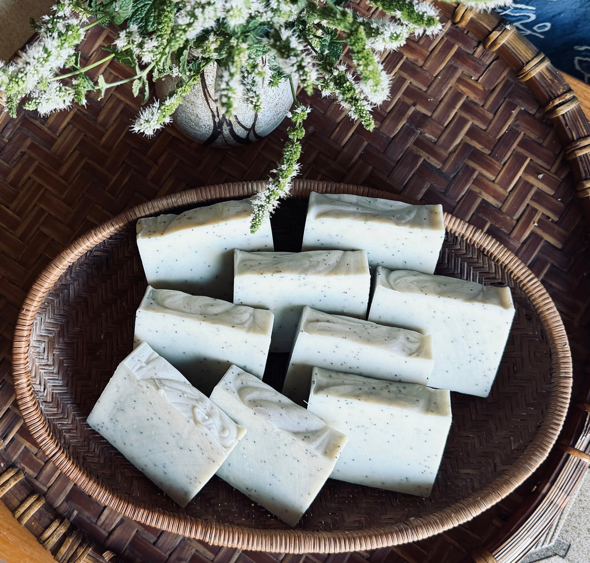 Several rectangular bars of pale green soap with small dark flecks are arranged in a woven oval basket on a larger wicker tray, with white flowers in the foreground.
