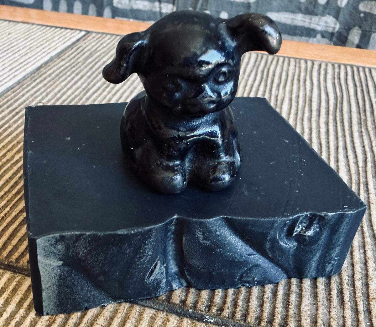 A small glossy black figurine of a seated dog or puppy with floppy ears, displayed on top of a matching black bar of charcoal soap against a striped woven mat background.