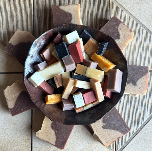 A top-down view of artisanal soap bars in various colors and marbled patterns, arranged in a star-like pattern around a dark ceramic bowl.