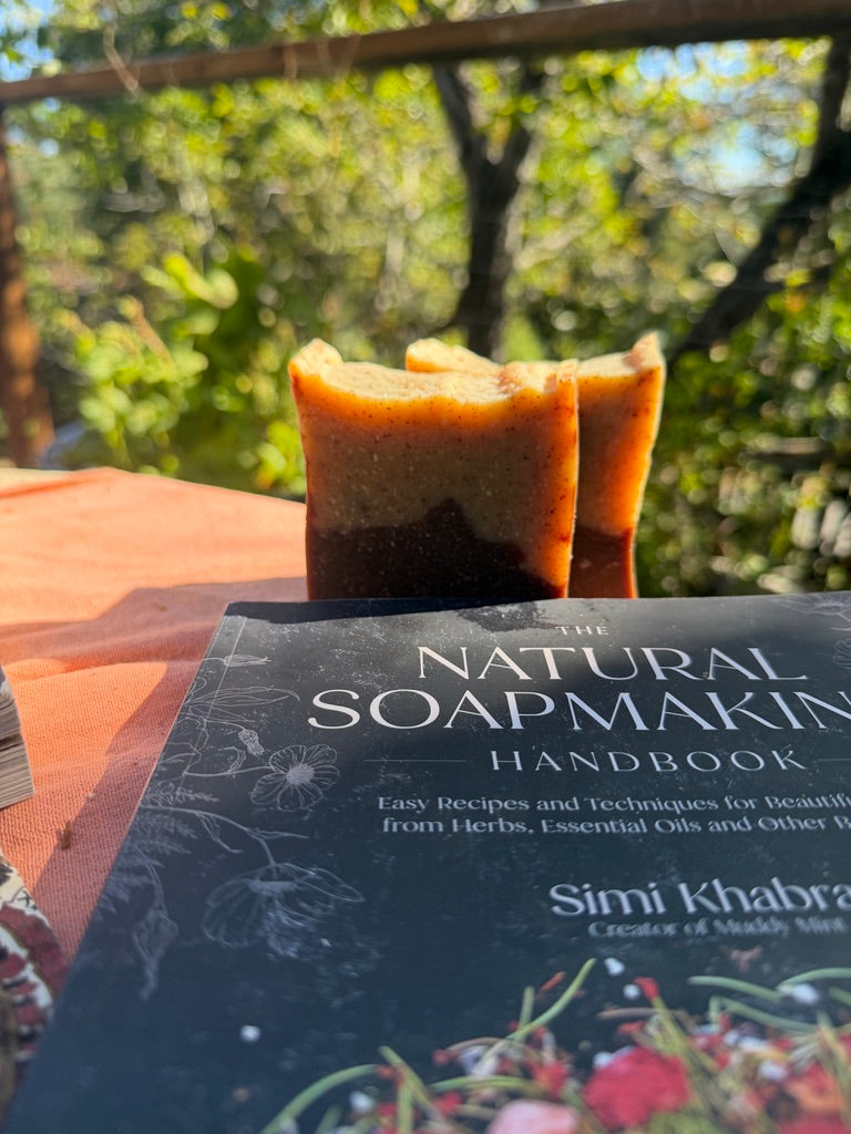 A book titled 'The Natural Soapmaking Handbook' by Simi Khabra lies on a table outdoors, with two handmade layered soap bars standing behind it against a blurred green garden background.