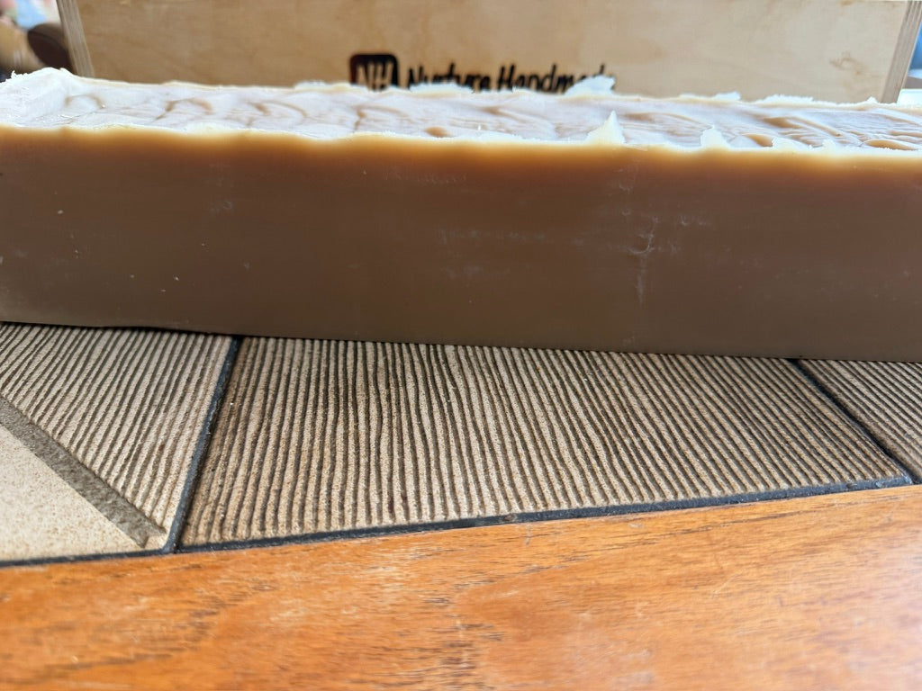 A thick rectangular bar of handmade soap in a warm tan-brown color, resting on a textured gray and beige tile table. A wooden soap mold with 'Nature Handmade' branding is visible in the background.