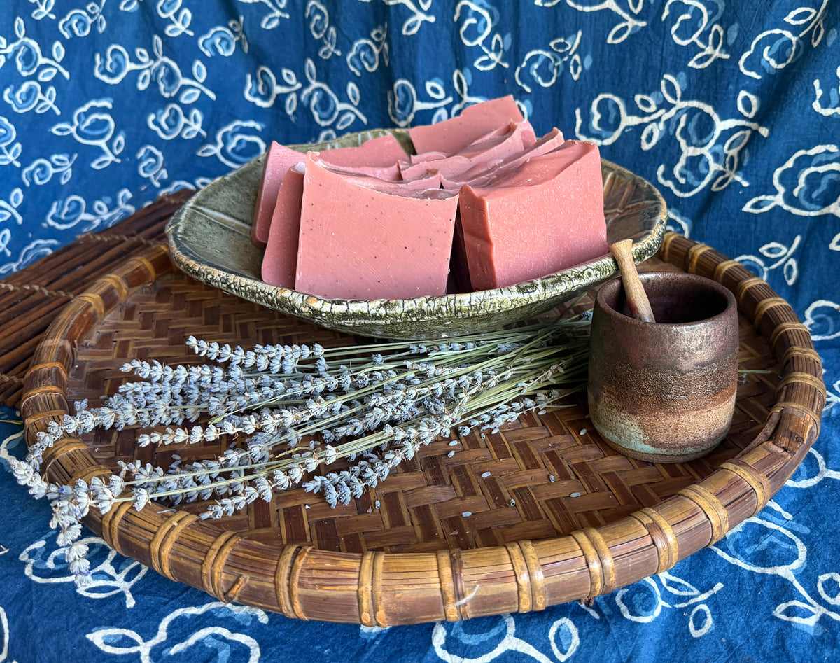 Pink rectangular bars of soap arranged in a small green ceramic dish on a round woven bamboo tray, with dried lavender sprigs and a small brown ceramic cup on a blue patterned fabric background.