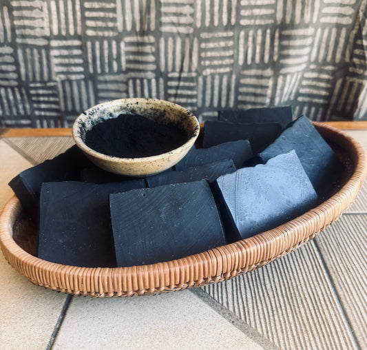 Activated charcoal blocks of soap arranged in a woven oval basket, with a small speckled ceramic bowl filled with activated charcoal set on a tiled surface against a patterned backdrop.