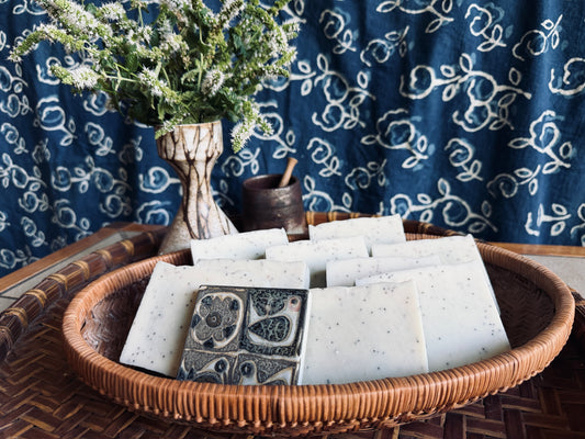 A woven wicker basket holds several rectangular handmade soap bars, including one with a dark floral pattern and others in a pale speckled tone. The basket sits on a woven surface in front of a blue patterned fabric backdrop, with a small vase of white flowers and a dark wooden tool behind it.
