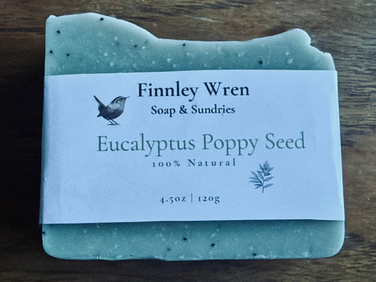 A rectangular bar of Finnley Wren Soap & Sundries 'Eucalyptus Poppy Seed' soap resting on a dark wooden surface. The soap is a pale sage green with dark speckles, and it has a white paper label wrapped around it.