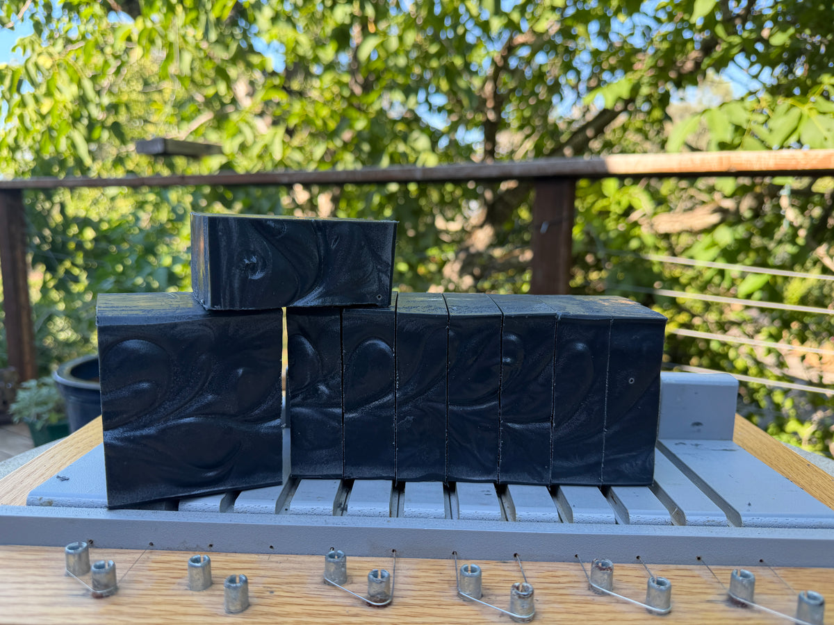 Several rectangular black soap bars with a swirled marbled pattern are stacked on a soap cutter with a garden and railing in the background.