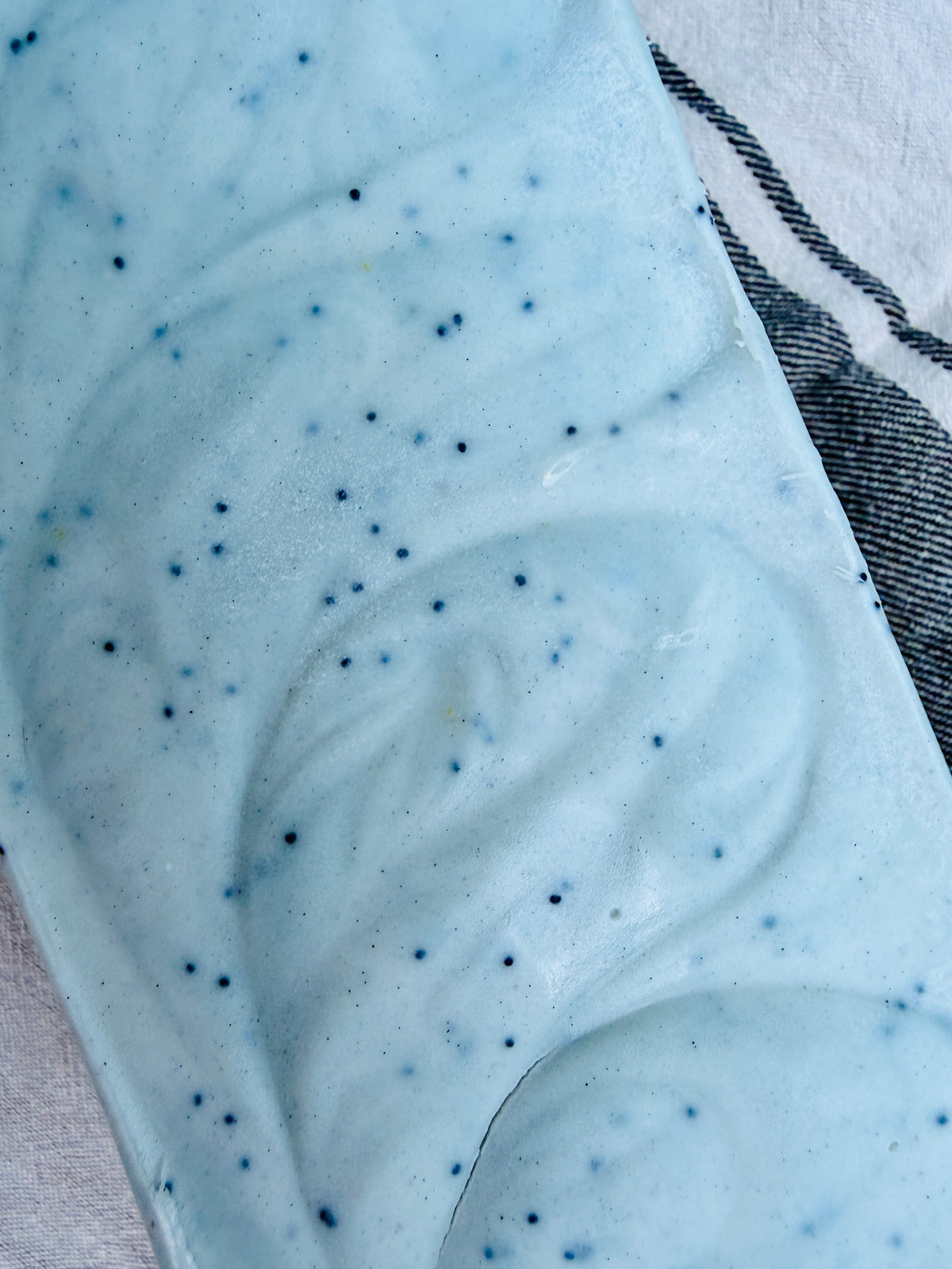A close-up of a light blue uncut loaf of handmade soap with small dark blue speckles, resting on a white fabric surface next to a gray striped cloth.