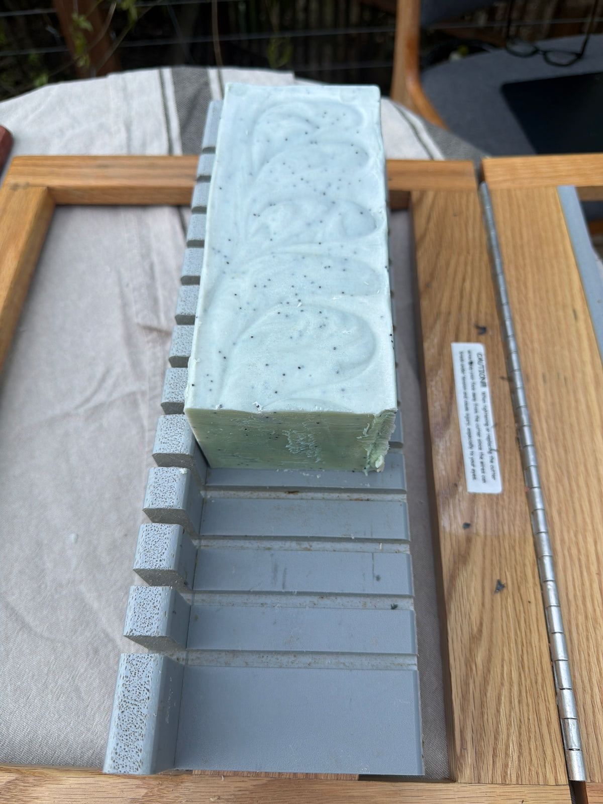 A rectangular bar of pale mint green soap with dark flecks, resting on a soap slicer.