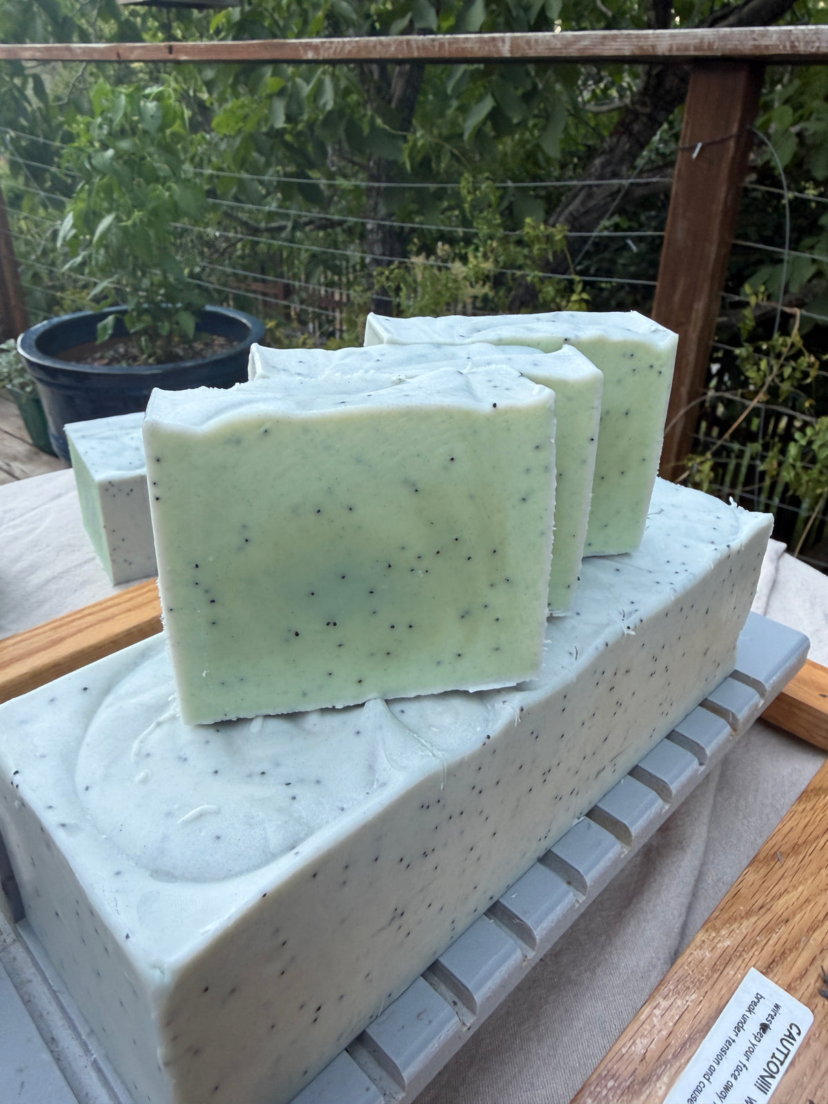 Three rectangular bars of pale mint green soap with dark speckles, displayed on top of a larger matching soap block outdoors on a wooden table with garden foliage in the background.