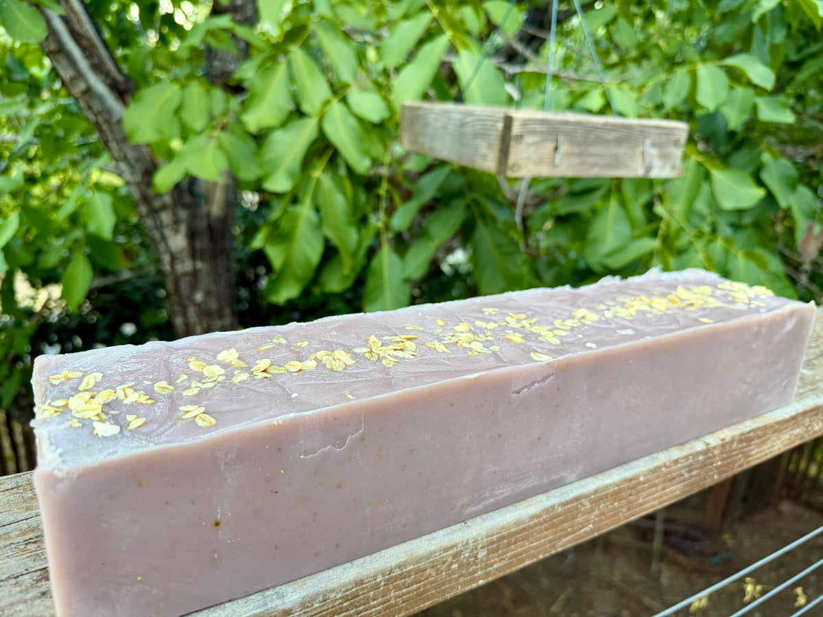 A large loaf of lavender-colored soap topped with dried oats, resting on a wooden surface outdoors with green foliage in the background.

