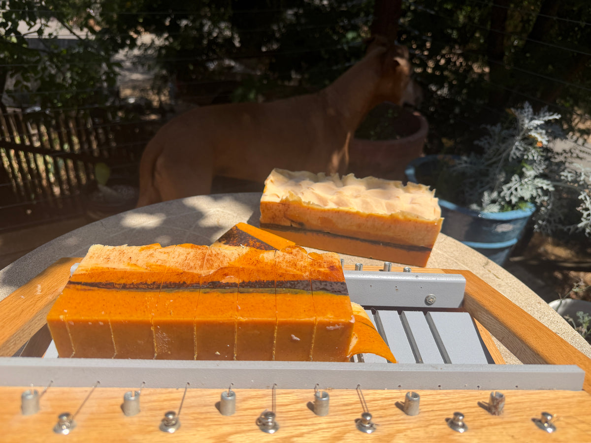 A close-up of a wooden and metal soap cutter in use, with two rectangular soap blocks positioned on the guide. A dog is visible in the background of an outdoor backyard setting with plants and fencing.


