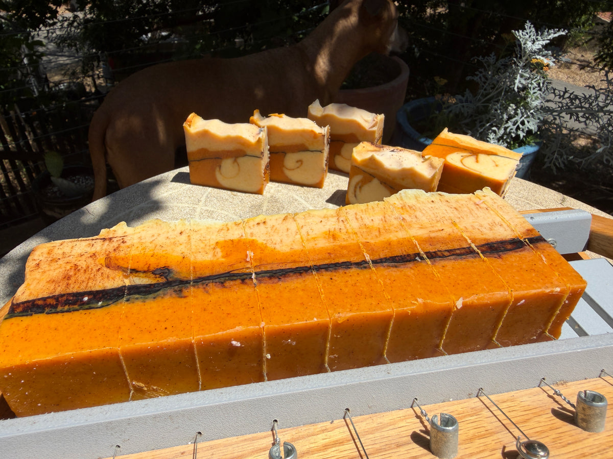 A large loaf of handmade soap with a glossy amber-orange finish and a dark center line, displayed outdoors on a soap cutter which sits on a table with several smaller soap bars behind it. A dog is visible in the background.

