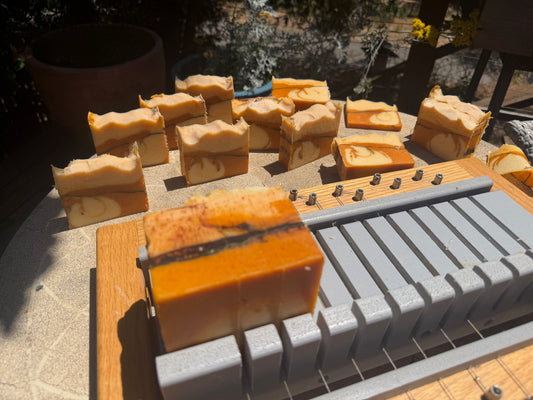 Handcrafted rectangular bars of soap in warm tan, orange, and cream swirls, displayed next to a wooden soap cutter in an outdoor setting.

