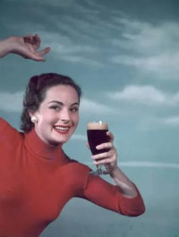 A smiling woman in a red turtleneck sweater holds a glass of dark beer against a painted blue sky background.